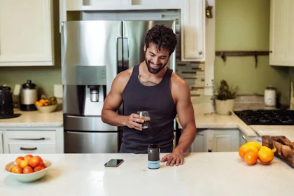 A man in a kitchen holding a glass of water, a supplement container on the counter.